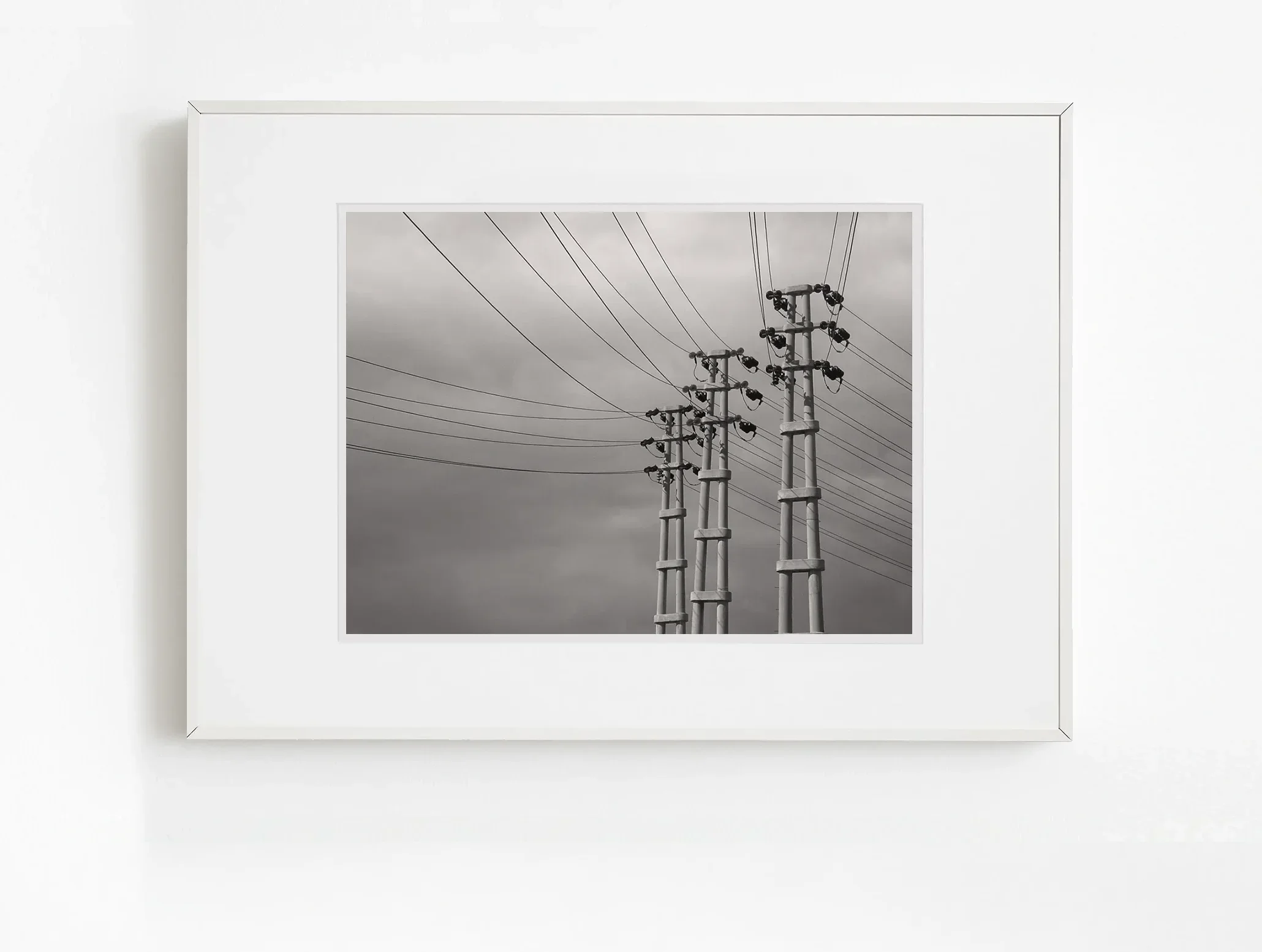 Bulutlu gökyüzü altında hizalanmış üç elektrik direği, yukarı doğru uzanan kablolarla minimal ve çağdaş siyah beyaz sanat fotoğrafı.